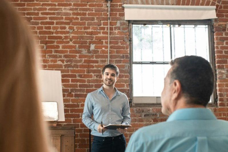 Young man presenting to a small group in a rustic brick interior room, holding a clipboard.